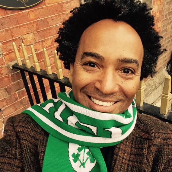 Thomas Glave, a black man smiling and wearing a green and white scarf and brown checked jacket