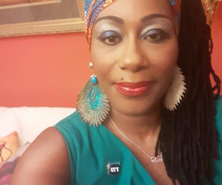 A person takes a selfie in a warmly lit room with red-orange walls. They wear a green sleeveless top, bold earrings, a vibrant headwrap, and striking makeup with blue eyeshadow and red lipstick. A pin reading "FIU" is visible on their top. Behind them is a white couch with pillows and a framed painting, adding to the cozy, expressive atmosphere.