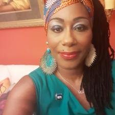 A person takes a selfie in a warmly lit room with red-orange walls. They wear a green sleeveless top, bold earrings, a vibrant headwrap, and striking makeup with blue eyeshadow and red lipstick. A pin reading "FIU" is visible on their top. Behind them is a white couch with pillows and a framed painting, adding to the cozy, expressive atmosphere.