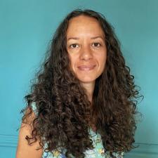 Photo of Jeda Pearl, who is a Scottish Jamaican woman with light-brown skin. She has dark brown curly hair, dark brown eyes and is smiling. She stands in front of a teal wall and is wearing a patterned dress with toucans and tropical leaves on it.