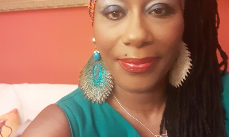 A person takes a selfie in a warmly lit room with red-orange walls. They wear a green sleeveless top, bold earrings, a vibrant headwrap, and striking makeup with blue eyeshadow and red lipstick. A pin reading "FIU" is visible on their top. Behind them is a white couch with pillows and a framed painting, adding to the cozy, expressive atmosphere.
