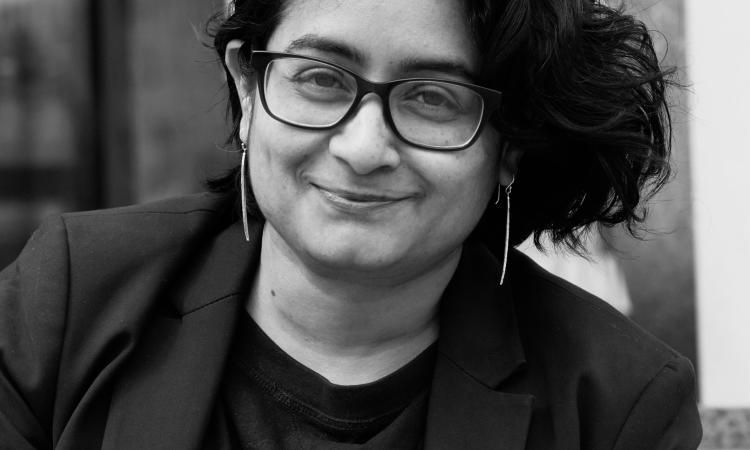 Black-and-white photo of a person with short, wavy hair and glasses, wearing a dark blazer and shirt, looking directly at the camera with a slight smile. The background includes a blurred sign and urban elements like steps.